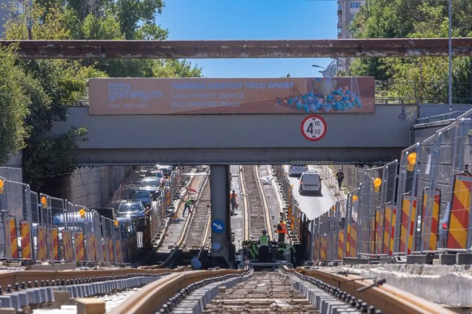 Șine de tramvai și balast se întind spre un pod de beton sub care circulă mașini pe un drum asfaltat.