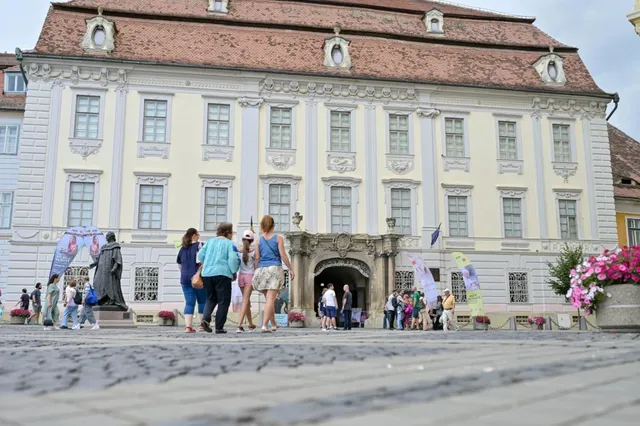 Oamenii stau pe o piață pavată în fața unei clădiri mari, cu fațadă deschisă la culoare și ferestre multiple, lângă o statuie și flori roz.