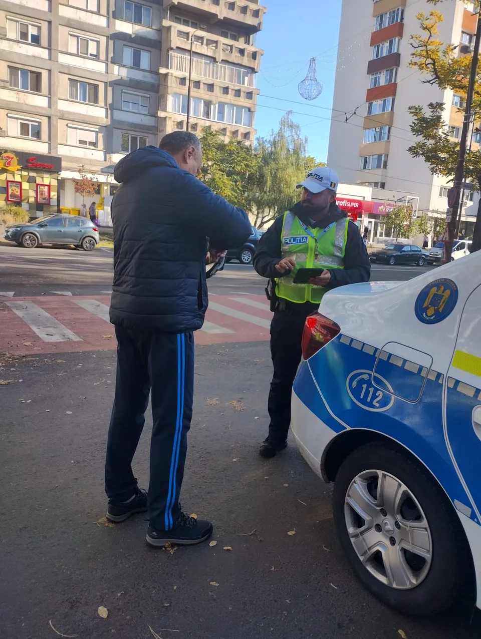 Un bărbat în haine închise stă în fața unui polițist care poartă vestă reflectorizantă și păstrează un vehicul alb cu marcaje albastre.