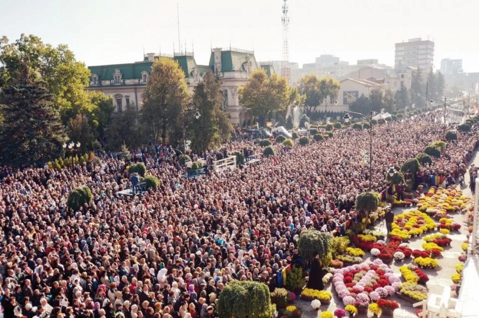 O mulțime mare de oameni este adunată pe o stradă lată lângă straturi de flori colorate și copaci decorați.