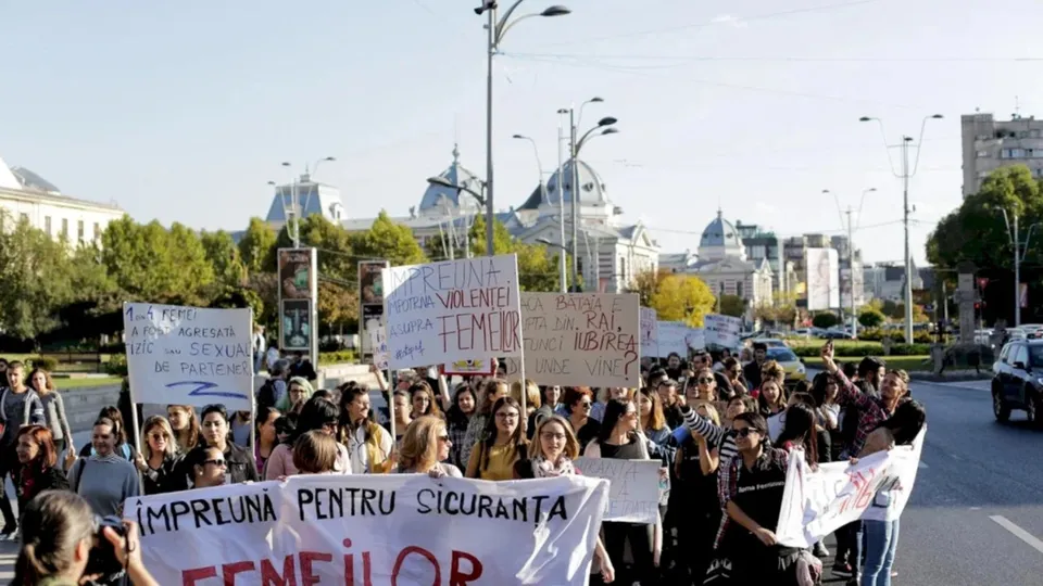 O mulțime de oameni merge pe un drum, purtând diverse panouri albe cu inscripții pe fundalul unor clădiri și copaci sub un cer luminos.