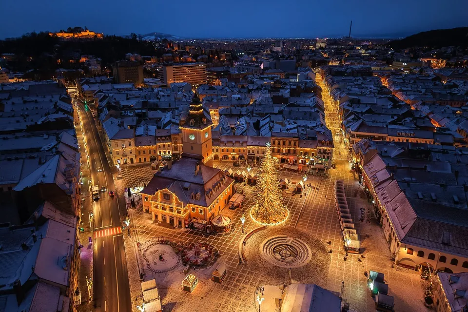 O vedere aeriană nocturnă a unui oraș cu clădiri iluminate, o piață pavată, un brad decorat și străzi cu mașini circulând.