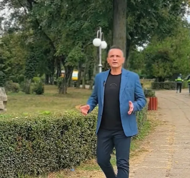 Un bărbat în sacou albastru și tricou negru stă pe o alee într-un parc, cu arbuști tunși și copaci în fundal. În depărtare se văd alte persoane.