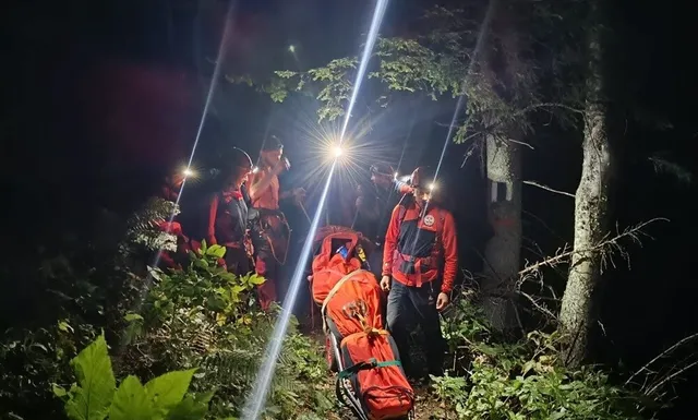 Patru persoane cu echipament de munte și lanterne porți într-o pădure întunecată, transportând un sac de salvare de culoare portocalie pe teren accidentat.