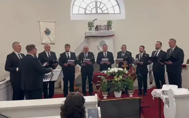 Un grup de bărbați în costume negre și cămași albe stau în rând, ținând partituri de muzică într-o încăpere cu banci de biserică.
