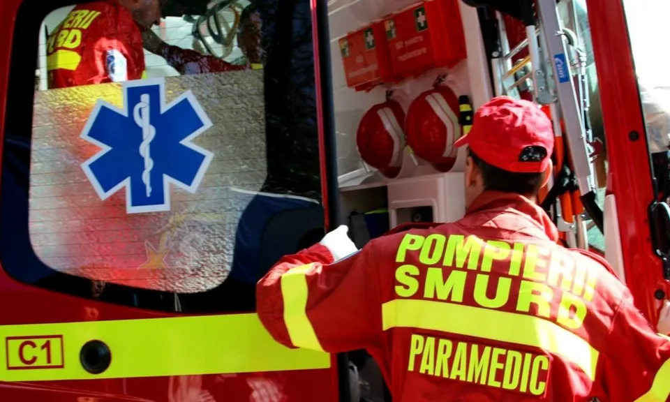 Un salvator cu uniformă roșie și inscripții galbene stă lângă un vehicul de urgență deschis, având echipamente medicale și căști în interior.