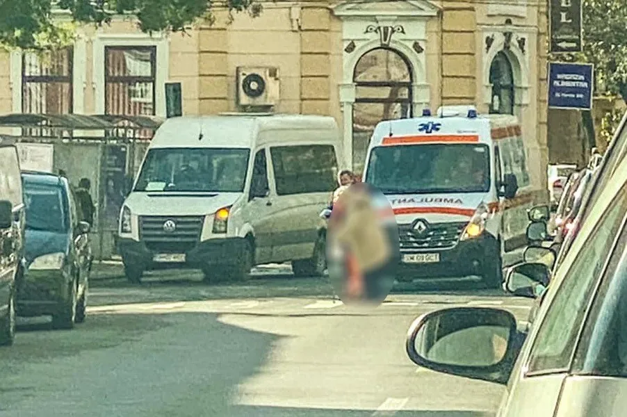 Un vehicul alb de tip van și o ambulanță sunt parcate pe o stradă lângă o clădire cu ferestre înalte și un aparat de aer condiționat.