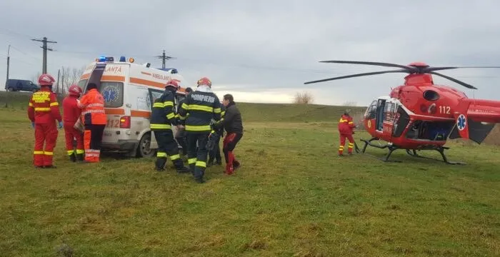Personal de intervenție în uniforme colorate stă lângă o ambulanță și un elicopter roșu pe un câmp de iarbă sub un cer înnorat.