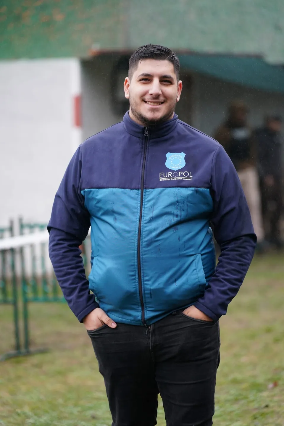 Un bărbat zâmbind poartă o jachetă albastră cu logo-ul Europol și pantaloni negri, stând cu mâinile în buzunare pe un fundal de iarbă și clădiri.