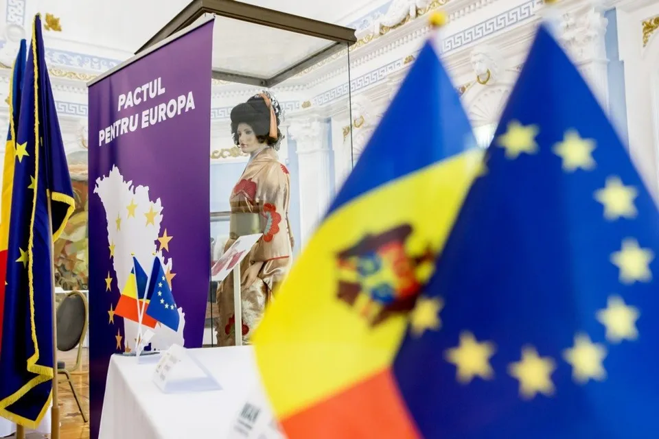 Steagurile Republicii Moldova și al Uniunii Europene sunt plasate în prim-plan, în fața unui panou violet și al unei statui într-o încăpere decorată.