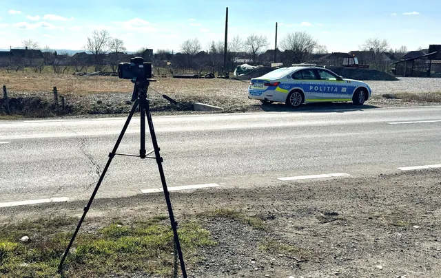 Un aparat foto montat pe un trepied negru este poziționat pe marginea unui drum asfaltat lângă o mașină de poliție albastră și albă.