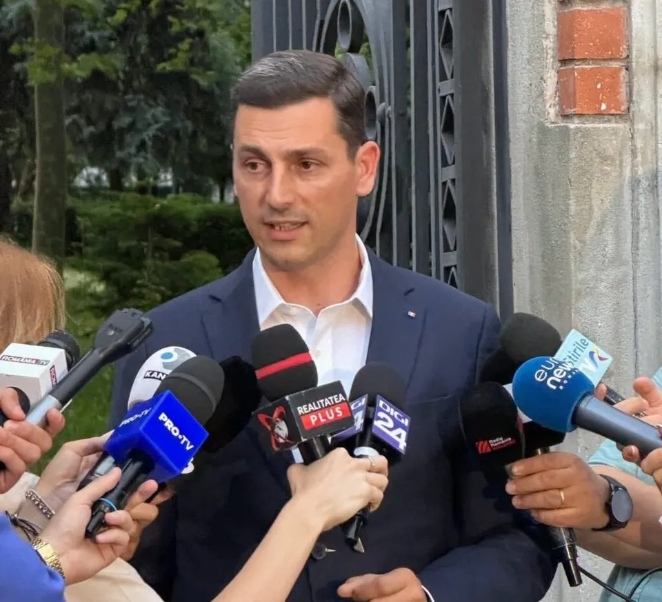 Un bărbat într-un costum albastru și cămașă albă vorbește în fața mai multor microfoane de la jurnaliști.