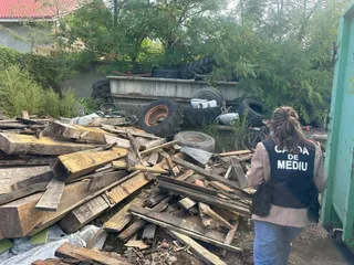 O persoană cu un vest vest de culoare închisă pe care scrie text alb stă în fața unui morman de lemn și a unor anvelope uzate.