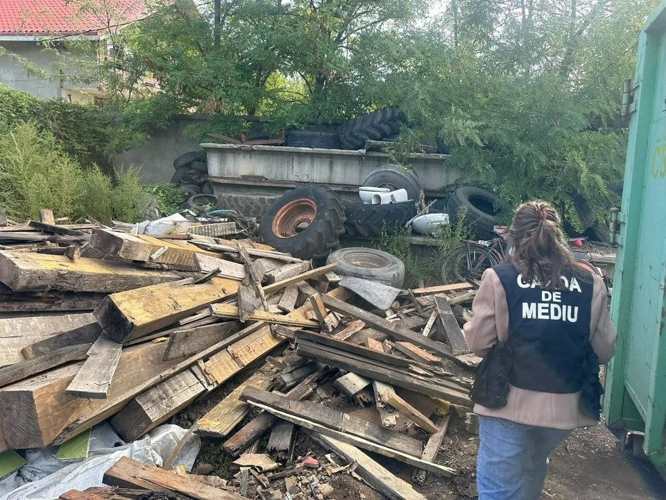 O persoană cu un vest vest de culoare închisă pe care scrie text alb stă în fața unui morman de lemn și a unor anvelope uzate.