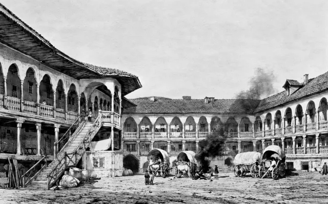 O curte interioară cu clădiri pe două etaje dispunând de arcade și balcoane, având căruțe cu acoperiș și oameni în centru.