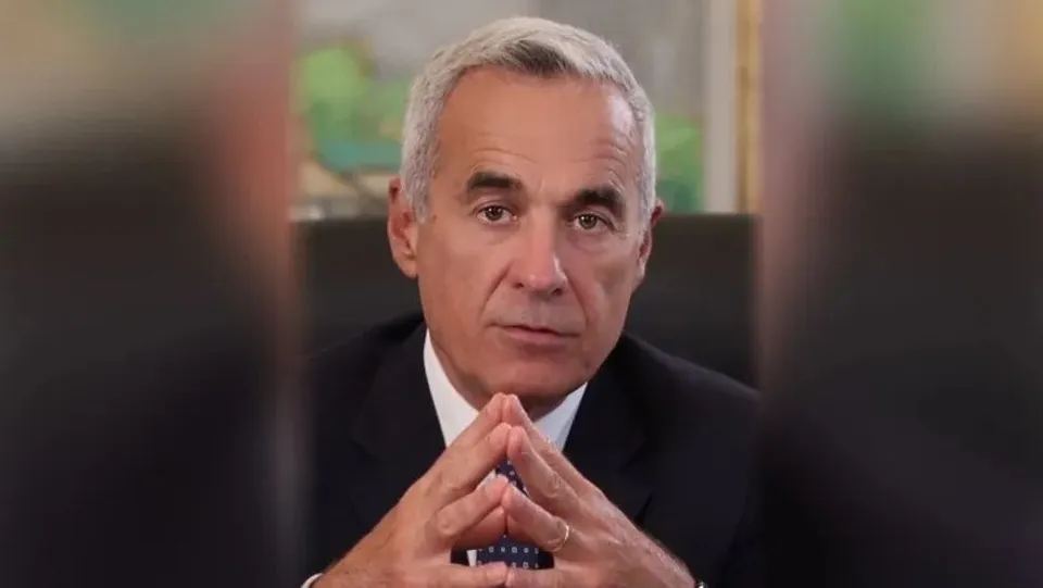 Un bărbat cu părul scurt și cărunt, purtând sacou închis și cămașă albă, stă cu mâinile împreunate sub bărbie.