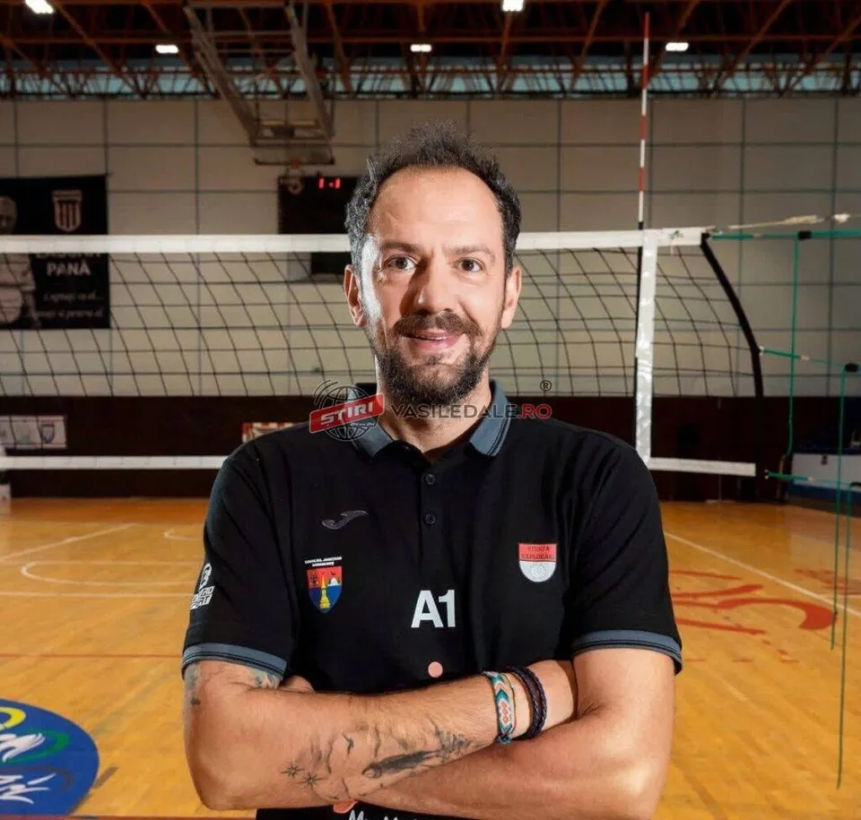 Un bărbat cu barbă și păr scurt stă cu brațele încrucișate într-o sală de sport cu un teren de volei în fundal.