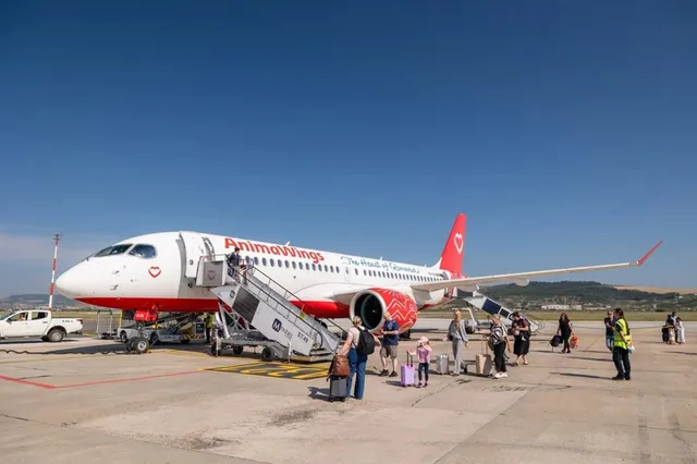 Un avion alb cu detalii roșii stă pe o pistă de aeroport, în timp ce pasageri cu bagaje stau în așteptare lângă o scară mobilă.