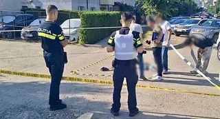 Unii polițiști în uniformă stau pe o stradă lângă o bandă de izolare galbenă, în timp ce un grup de persoane stă în grup în fundal.
