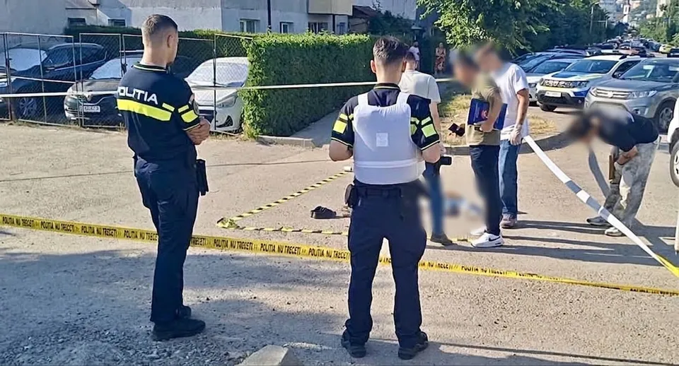Unii polițiști în uniformă stau pe o stradă lângă o bandă de izolare galbenă, în timp ce un grup de persoane stă în grup în fundal.
