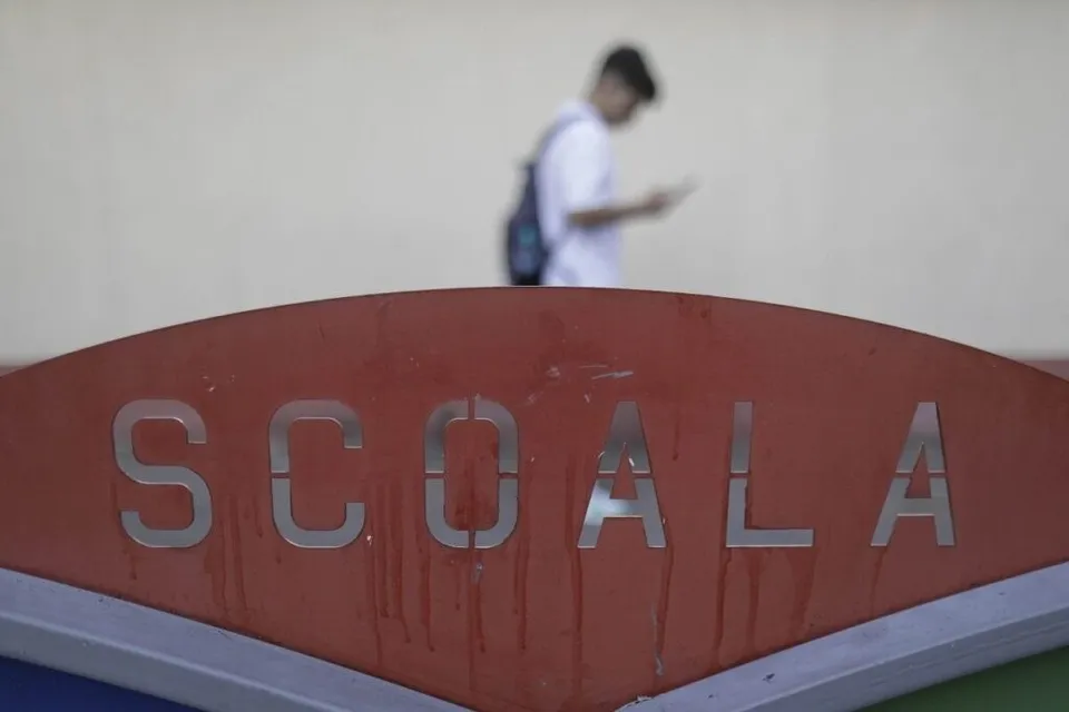 Un semn roșu cu textul metalic SCOALA este în prim-plan, în timp ce un elev cu rucsac stă în fundal pe un fundal deschis.