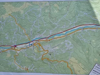 O hartă topografică detaliată prezintă linii de contur, drumuri colorate și zone verzi pe un fundal de relief montan.