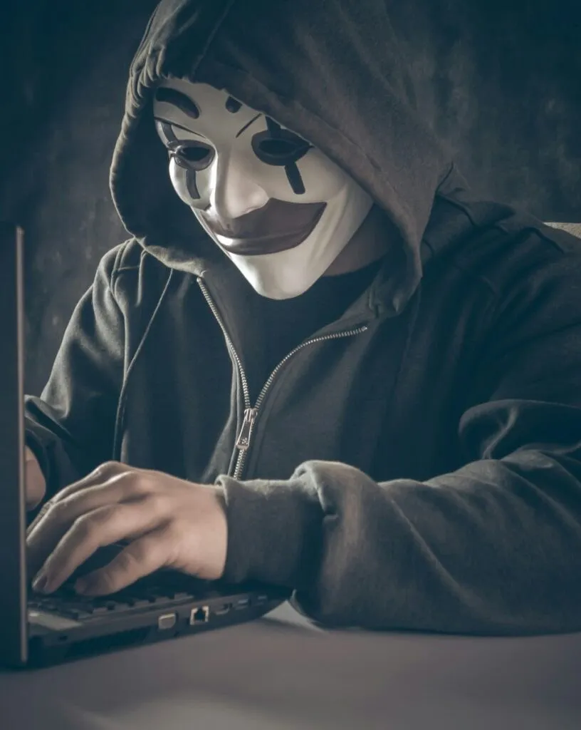 O persoană cu o mască de clovn și o capucină închisă tastează pe o laptop într-un mediu întunecat.