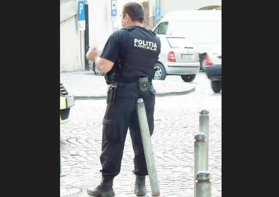 Un agent de poliție locală stă cu spatele, purtând un tricou negru cu inscripție albă și pantaloni închizi, lângă niște stâlpi metalici pe un trotuar.