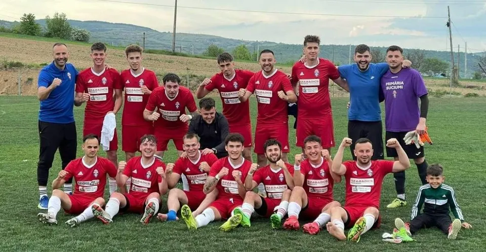Un grup de fotbaliști în echipamente roșii și albastre pozează pe un teren de fotbal verde sub un cer deschis.