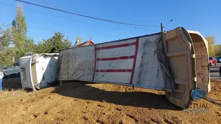 Un autotren cu o remorcă mare, de culoare deschisă cu dungi roșii, este răsturnat pe o suprafață de pământ lângă un drum.
