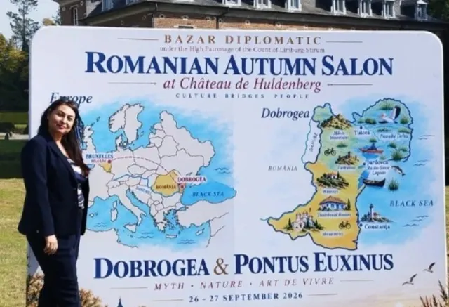 O femeie cu părul negru și o jachetă închisă la culoare stă lângă un panou informativ alb cu hărți și text colorat.