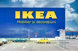 Un magazin cu un panou albastru mare pe care scrie IKEA și Mobilier și decorațiuni, aflat lângă un parc de parcare cu mașini colorate.