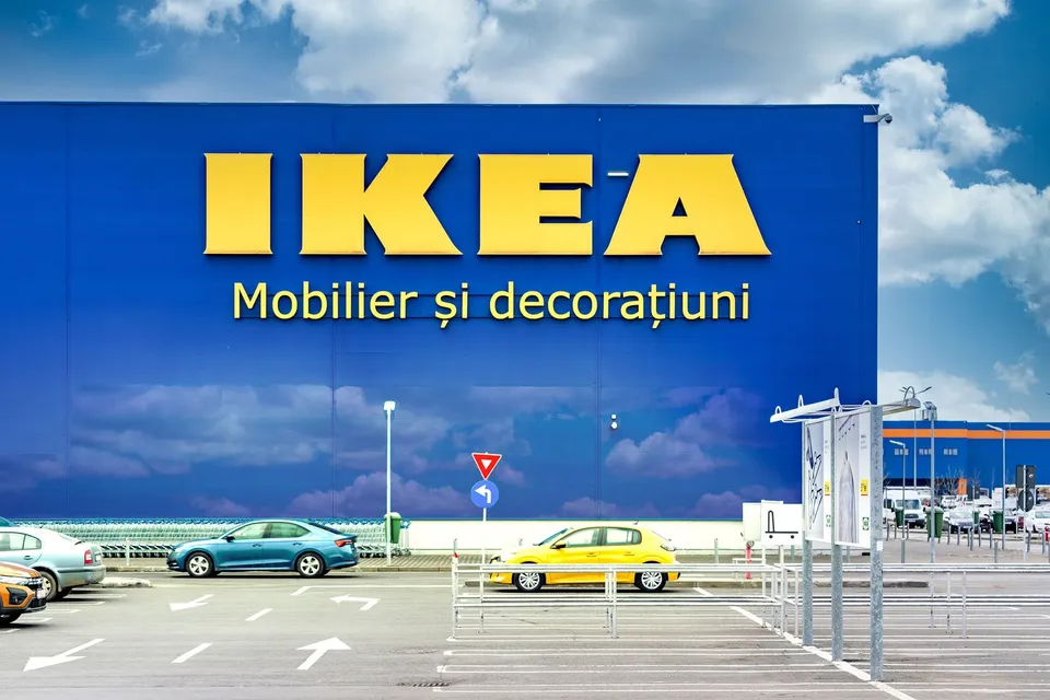 Un magazin cu un panou albastru mare pe care scrie IKEA și Mobilier și decorațiuni, aflat lângă un parc de parcare cu mașini colorate.