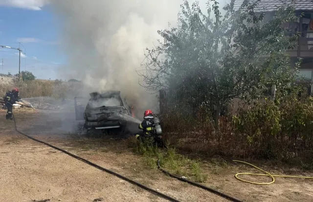 Un autoturism ars este înconjurat de fum dens, în timp ce un pompier în echipament de protecție se află lângă acesta pe un teren neasfaltat.