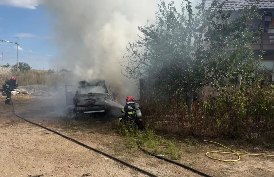 Un autoturism ars este înconjurat de fum dens, în timp ce un pompier în echipament de protecție se află lângă acesta pe un teren neasfaltat.
