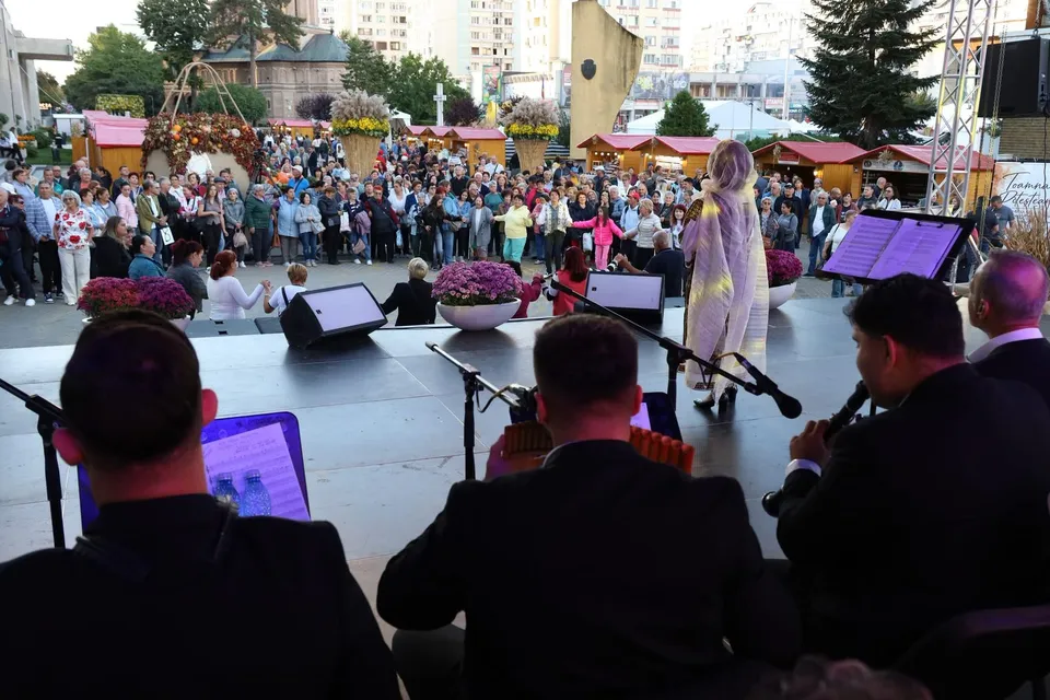 Din perspectiva unei scene, câțiva bărbați în haine negre privesc spre o mulțime de oameni adunată în fața unor tarabe de lemn.
