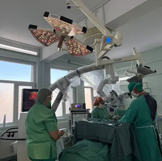Trei cadre medicale în uniforme verzi, cu măști, într-o sală de operație, în jurul unei mese de operație, cu echipament medical modern.