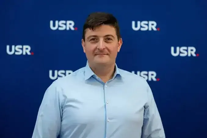 Un bărbat cu părul scurt și cămașă albastră deschisă stă în fața unui fundal albastru pe care sunt imprimate mai multe logo-uri albe și roșii.