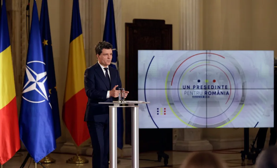 Un bărbat într-un costum închis la culoare stă în spatele unui podium lângă câteva steaguri și un ecran mare cu grafică circulară.