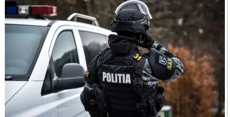 Un polițist cu echipament tactice negru și cască stă lângă un vehicul alb cu girofaruri.
