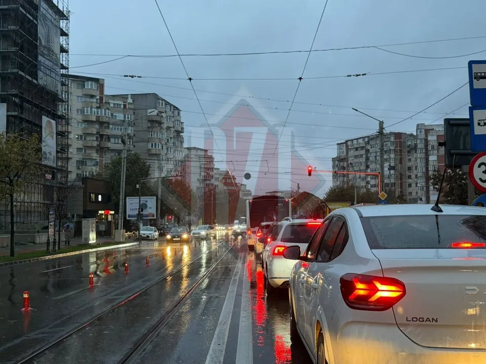 Imaginea prezintă un traffic auto intens pe o șosea ușor udă, pe un fundal cityscape într-o zi înnorată și ploioasă, cu mașini albe și gri în prim plan.