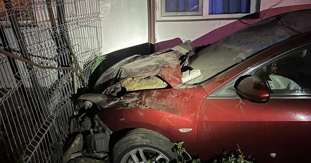 Un autoturism roșu cu partea frontală zdrobită este poziționat lângă un gard metalic și o clădire.