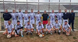 O echipă de fotbaliști stă în poziții diverse pe un teren de fotbal, în fața unei porți de fotbal.