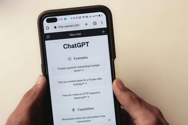O pereche de mâni ține un smartphone afișând interfața aplicației ChatGPT, cu un meniu de exempluri și capacități pe ecran.
