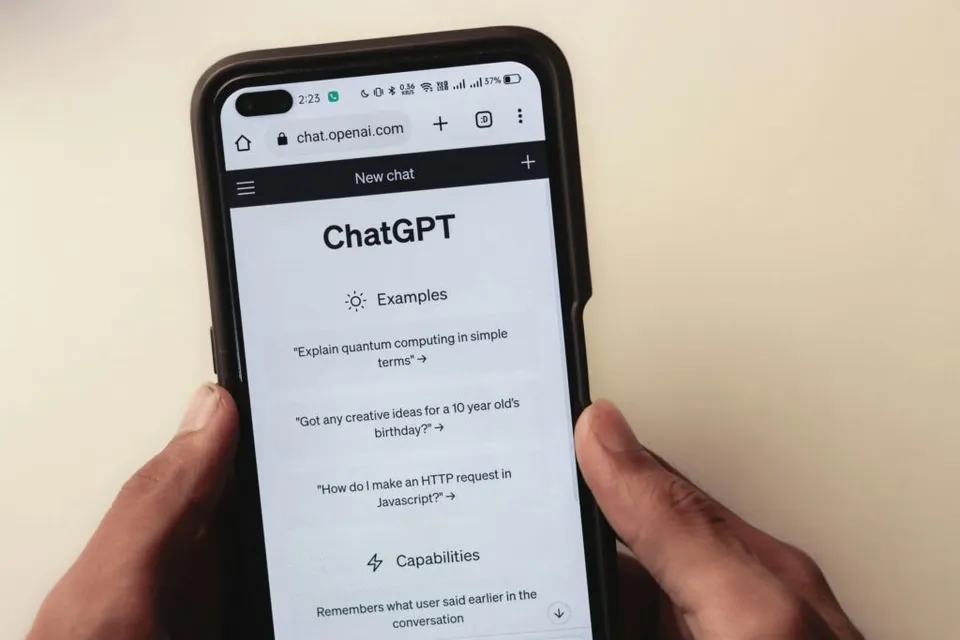 O pereche de mâni ține un smartphone afișând interfața aplicației ChatGPT, cu un meniu de exempluri și capacități pe ecran.