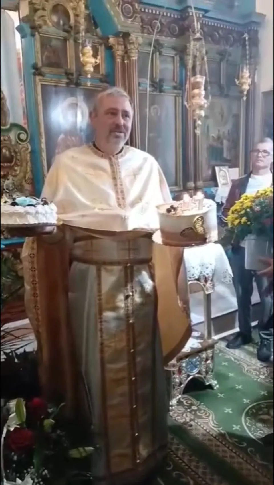 Un bărbat cu barbă poartă haine liturgice de culoare deschisă în fața unui iconostas decorat cu coloane și picturi religioase.