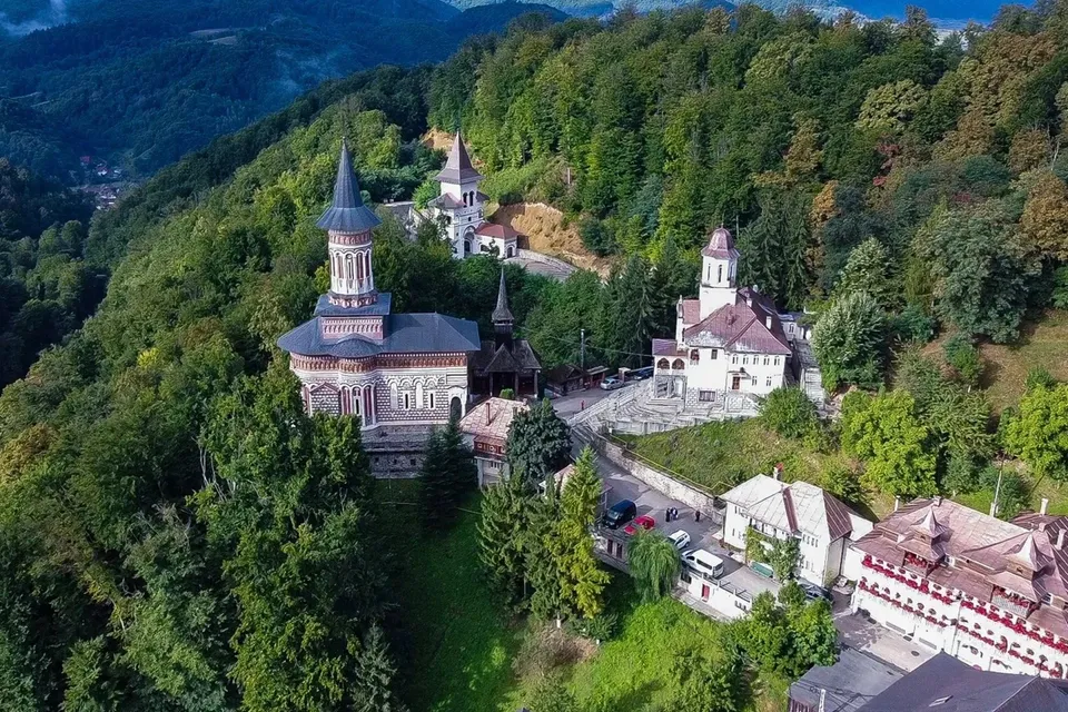 O vedere aeriană prezintă un complex de clădiri cu acoperișuri înclinate și turnuri înalte, înconjurat de o pădure densă de copaci verzi pe un deal.