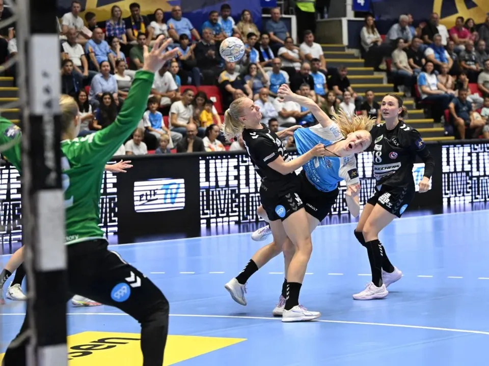 Câteva jucătoare de handbal în echipamente negre și albastre se află pe un teren de sport în fața unui public numeros.
