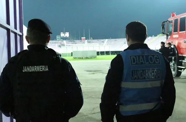 Doi bărbați în uniformă neagră cu inscripții de tip vestă sunt priviți din spate, privind spre un teren de fotbal iluminat noaptea.