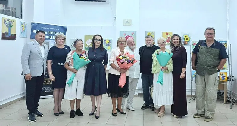 Un grup de zece persoane, bărbați și femei, stau în picioare într-o sală de expoziție, cu tablouri pe pereți și un banner albastru în fundal.